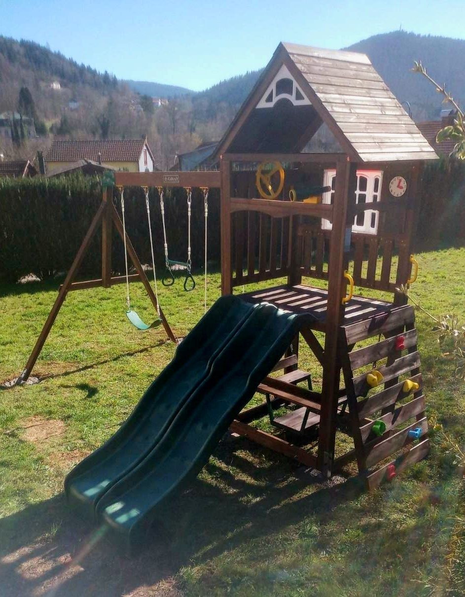 A wooden playhouse with a slide and swings in a grassy yard.
