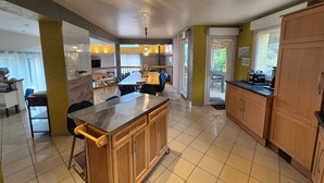 A modern kitchen with wooden cabinets, a stainless steel island, and a dining area with a large table and chairs. - Le Coeur des Vosges -  Jacuzzi disponible toute l'année (Cornimont)