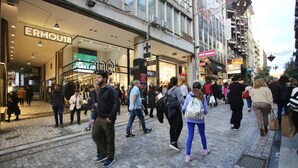 Una calle urbana concurrida con peatones y comercios, incluyendo letreros visibles para 'ERMOU18' y 'planet'.