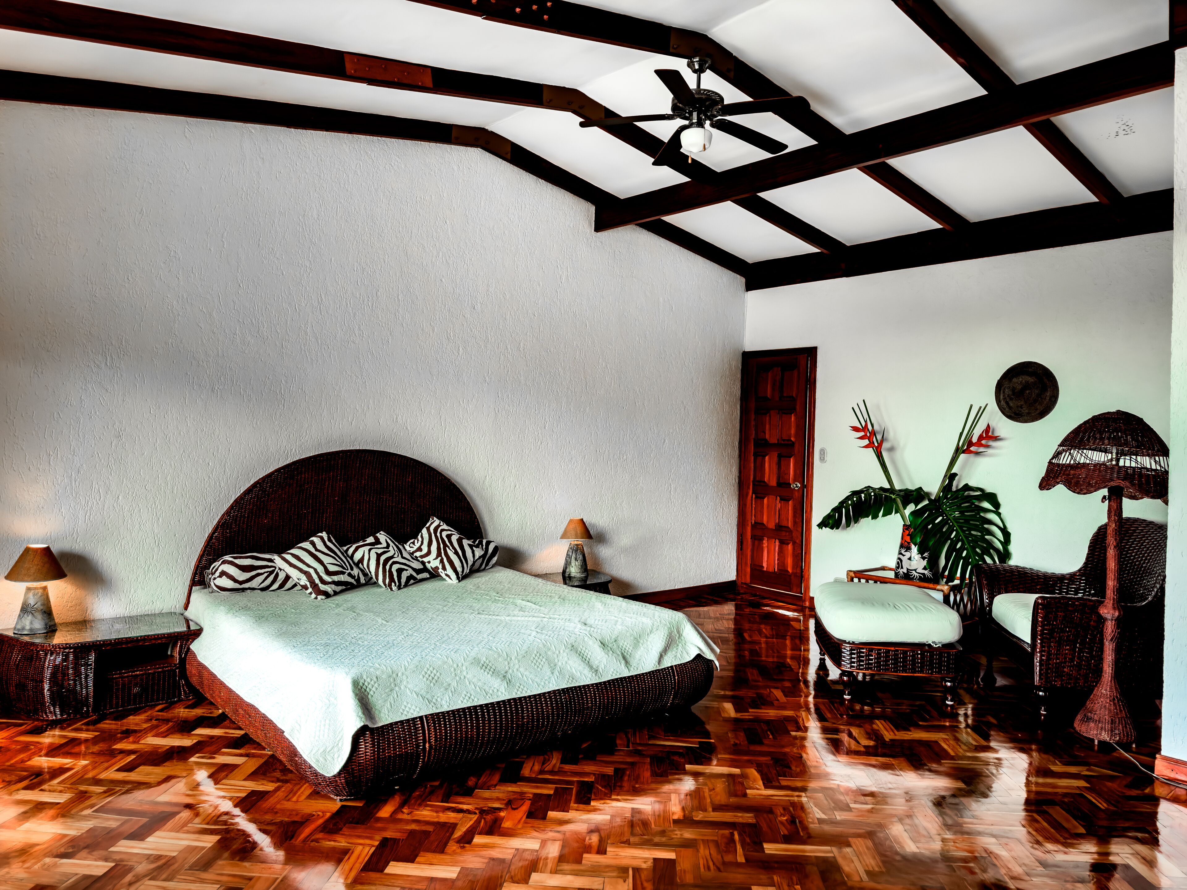 Ein Schlafzimmer mit Holzdielenmuster-Boden, einem Bett mit Zebra-Kissen und einem Deckenventilator, sowie einer an der Wand befestigten Pflanze.