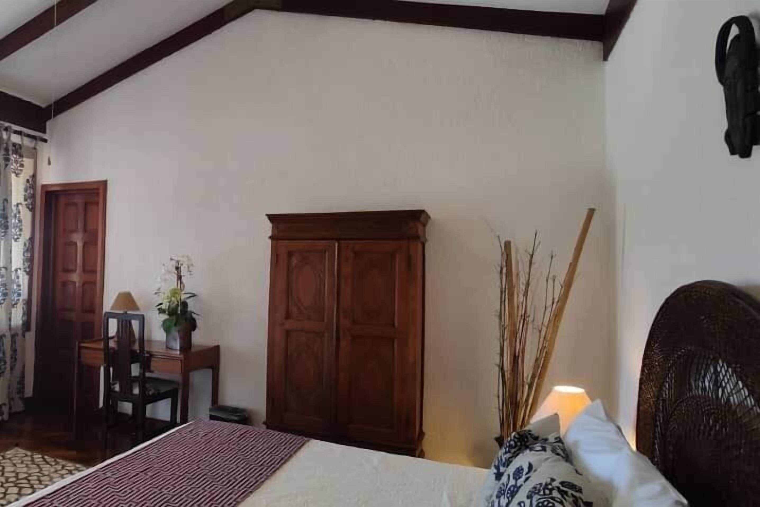 A bedroom with a bed, wooden wardrobe, desk with chair, and a lamp.