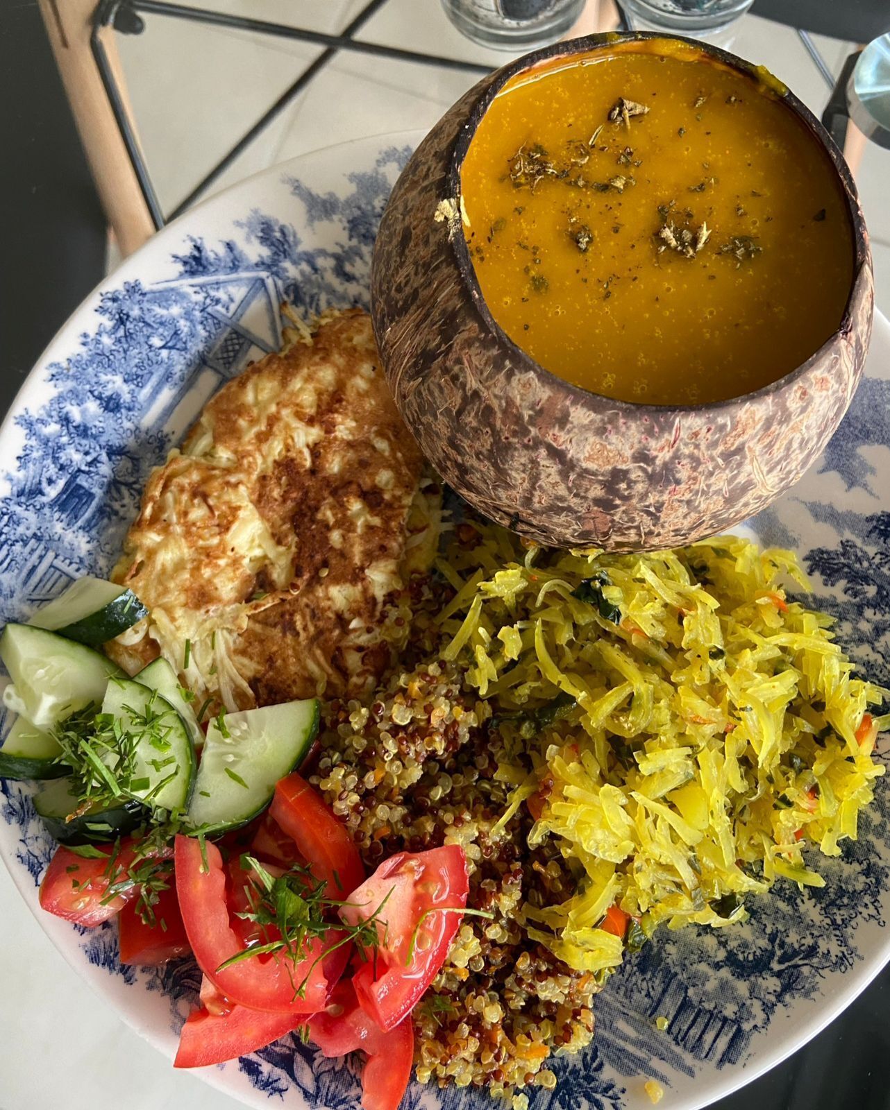 A plate with a bowl of soup, a piece of grilled chicken, a salad with cucumbers, tomatoes, and herbs, and a serving of yellow rice.