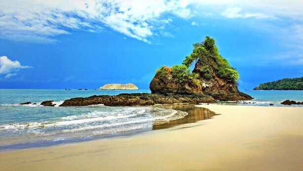 A beach with a large rock formation covered in greenery, clear blue water, and a sandy shore. - Canal Grande Lodge (Piedades)