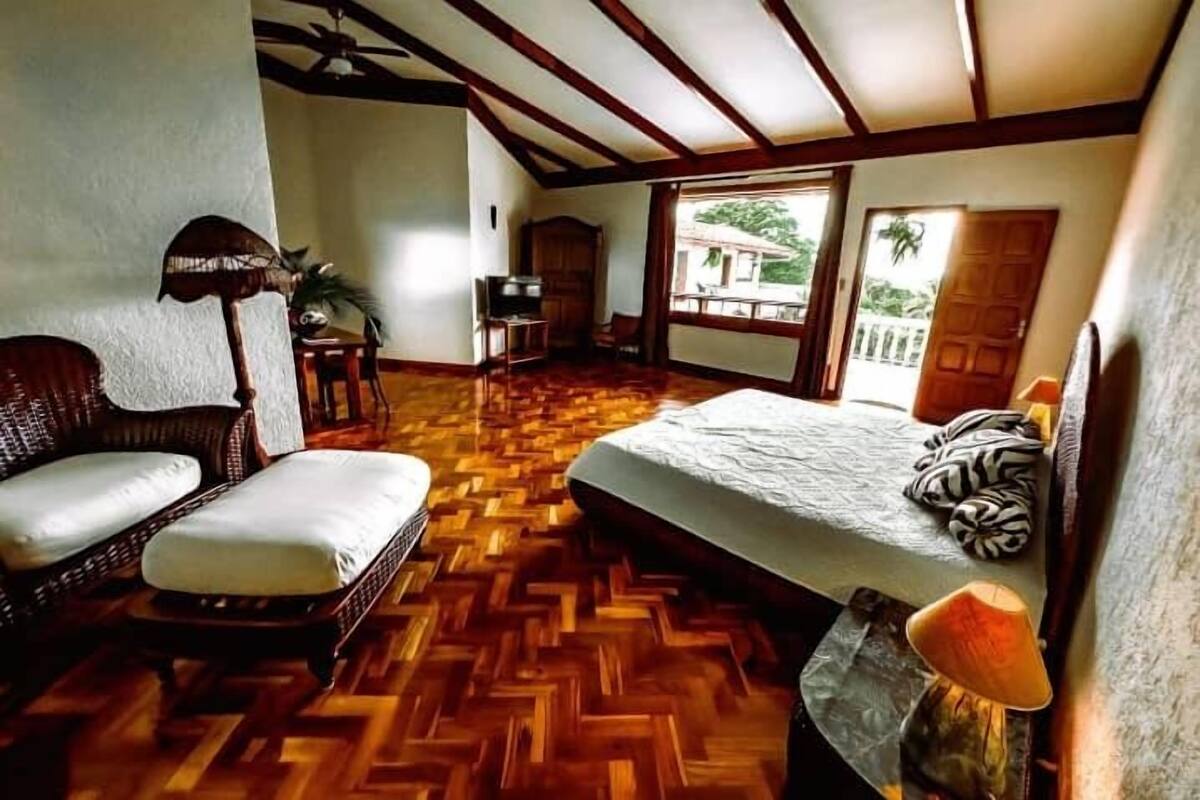 A room with a wooden herringbone floor, a bed with a white bedspread, a wicker sofa, a small table, and a ceiling fan.