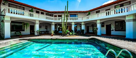 Zona de piscina con una piscina azul cristalina, rodeada por un edificio blanco con tejados de tejas rojas y varios balcones.