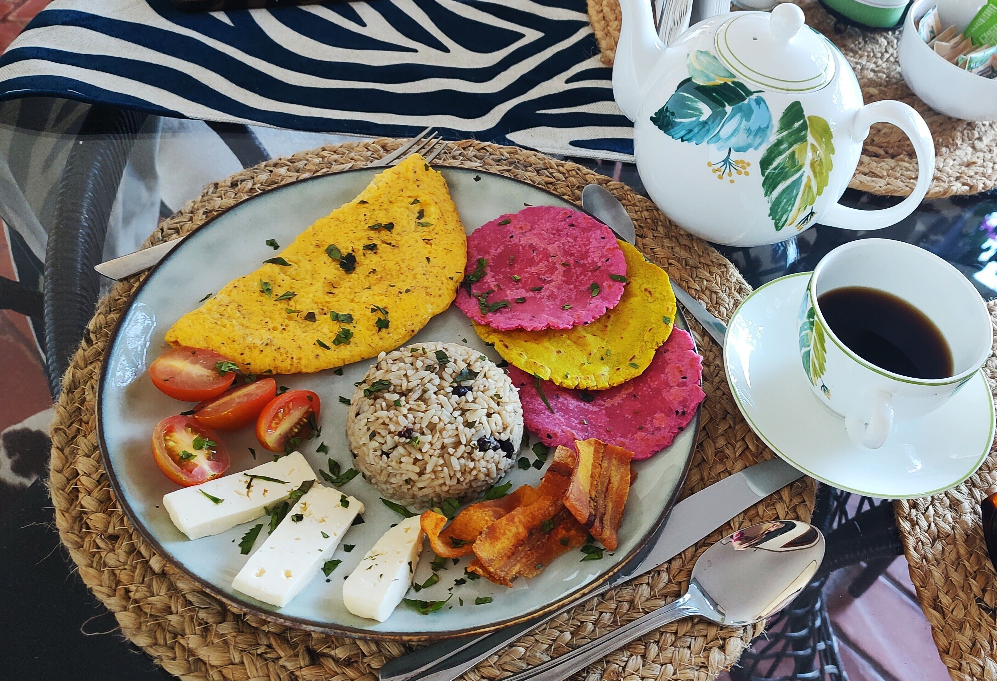 A breakfast plate with a variety of foods, a cup of coffee, and a teapot on a woven placemat.