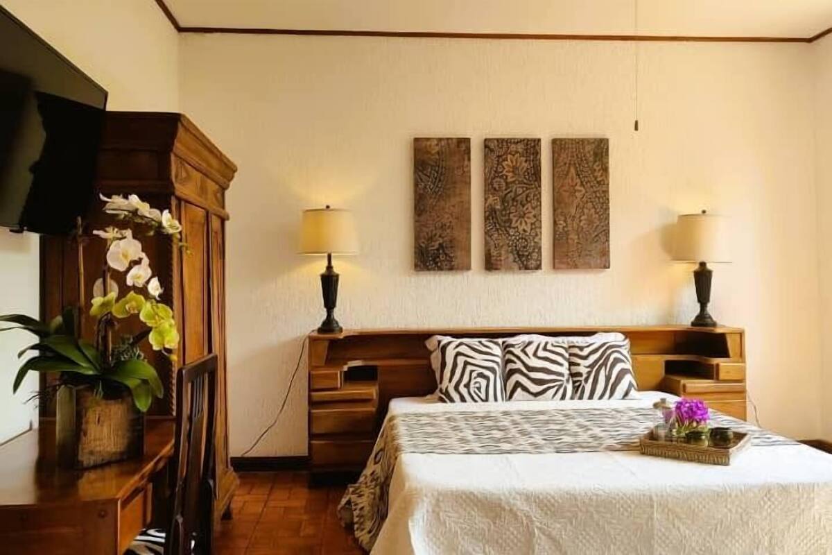 A bedroom with a bed, two wall-mounted lamps, a wooden nightstand, a dresser, and a potted plant.