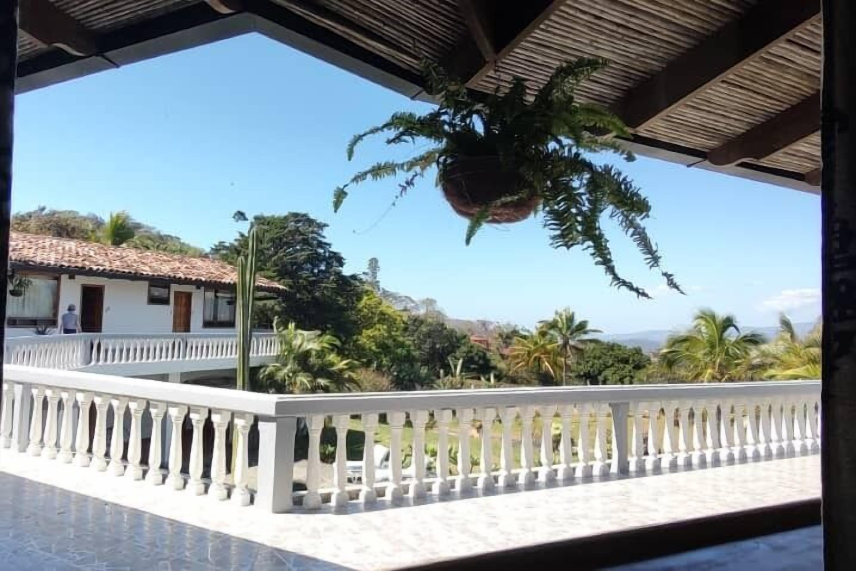 Área externa coberta com um parapeito branco, planta pendente e vista para um edifício com telhado de azulejos.