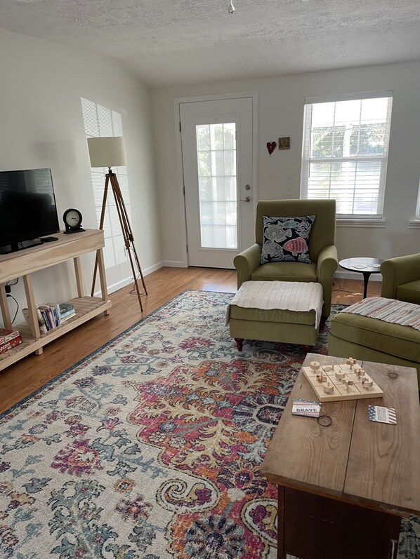A living room with a green armchair, a wooden coffee table with chess pieces, a television on a stand, a floor lamp, and a patterned rug. - Walk to Old St Andrews from cozy bungalow on Lake Huntington: The Crow’s Nest (Panama City)