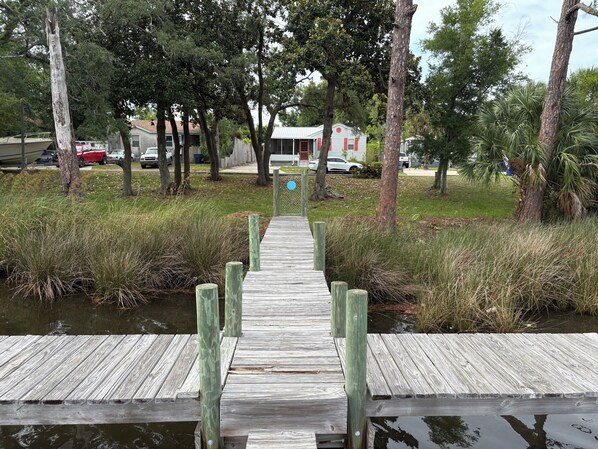 En träbrygga som leder fram till ett hus med en röd dörr, omgivet av träd och en gräsyta.