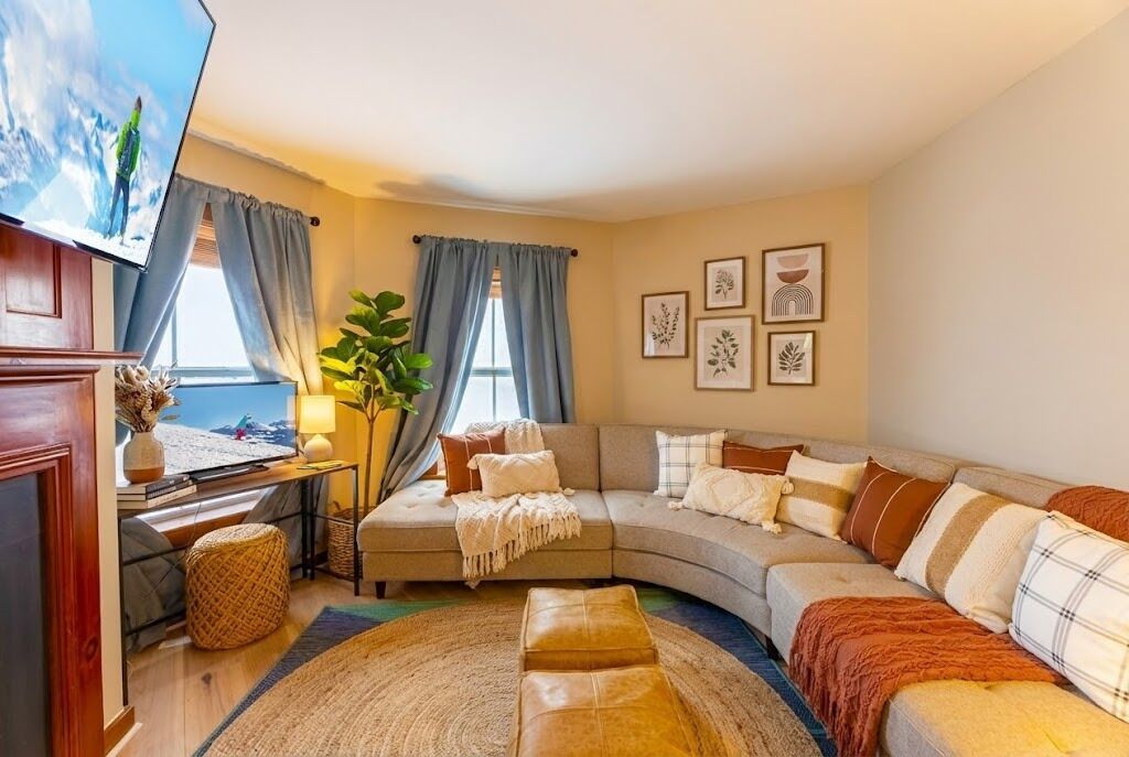 A cozy living room with a curved sofa, a flat-screen TV, and a large window with curtains.