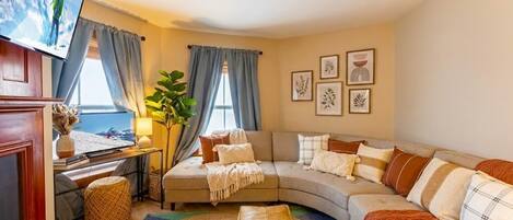 A cozy living room with a curved sofa, a flat-screen TV, and a large window with curtains.