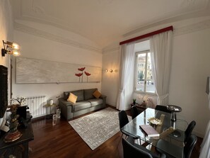 A modern living room with a leather sofa, a glass dining table, and a view of the street through a window with white curtains. - Your home in the center of Rome (Roma)