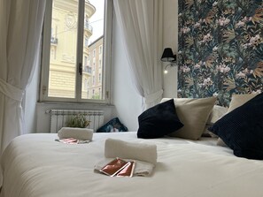 Una cama bien hecha con sábanas blancas, una pared con estampado floral y vistas a edificios a través de una ventana con cortinas blancas.