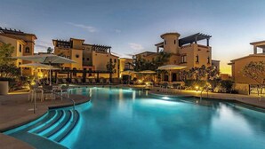 Een zwembad met een golvend ontwerp, omringd door ligbedden en parasols, met gebouwen in mediterrane bouwstijl op de achtergrond.