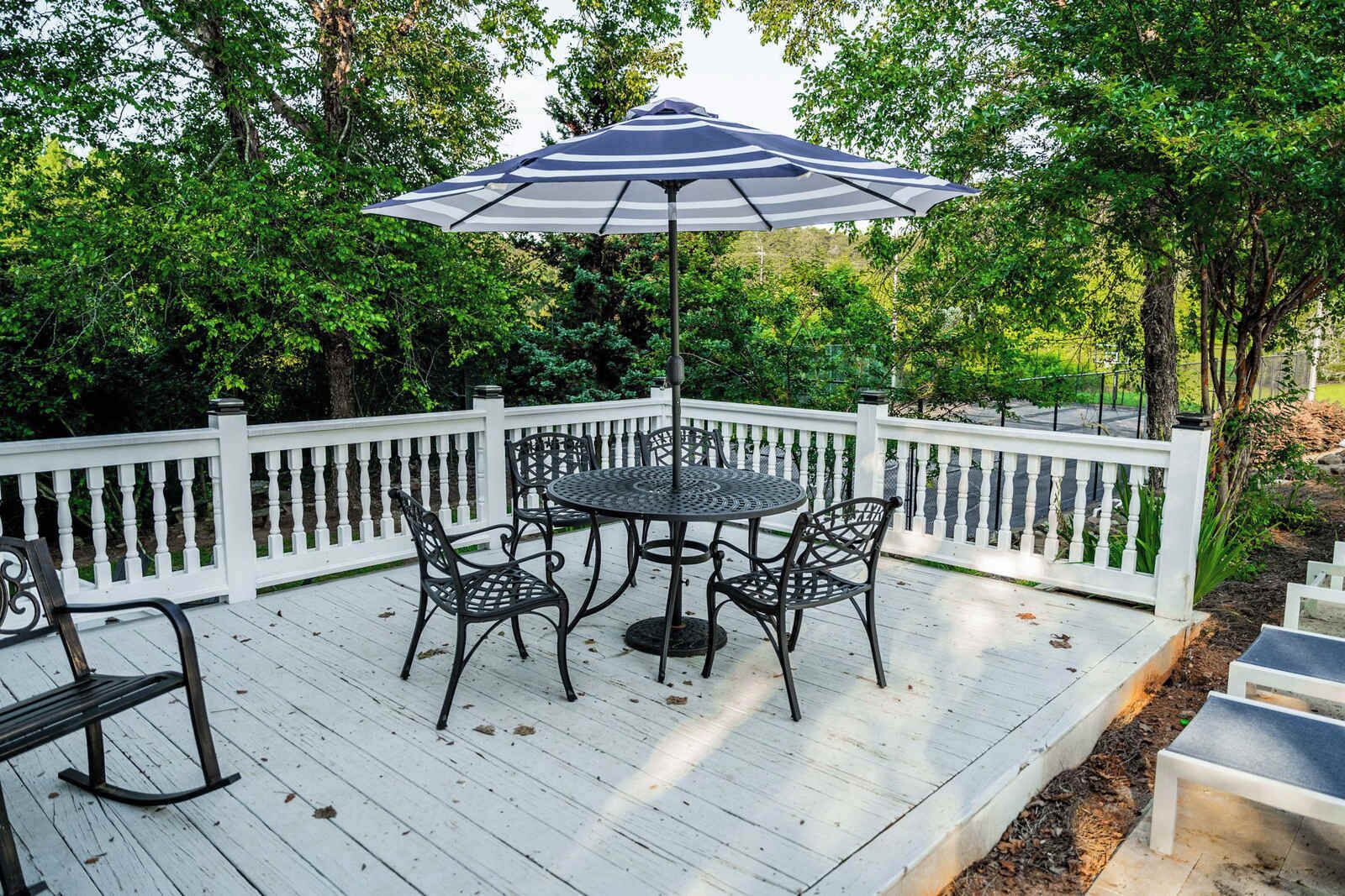 A deck with a round table and chairs, a striped umbrella, and a white picket fence.