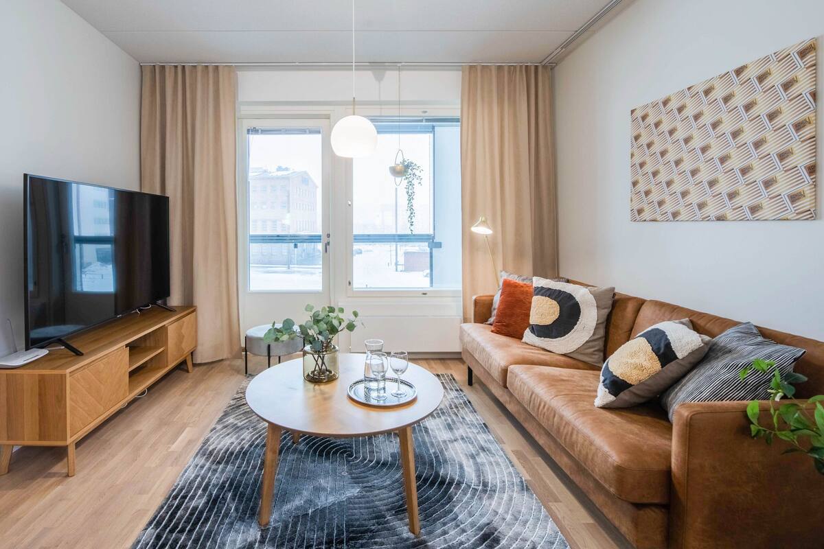 A modern living room with a flat-screen TV, wooden furniture, a round coffee table, and a large window with curtains.