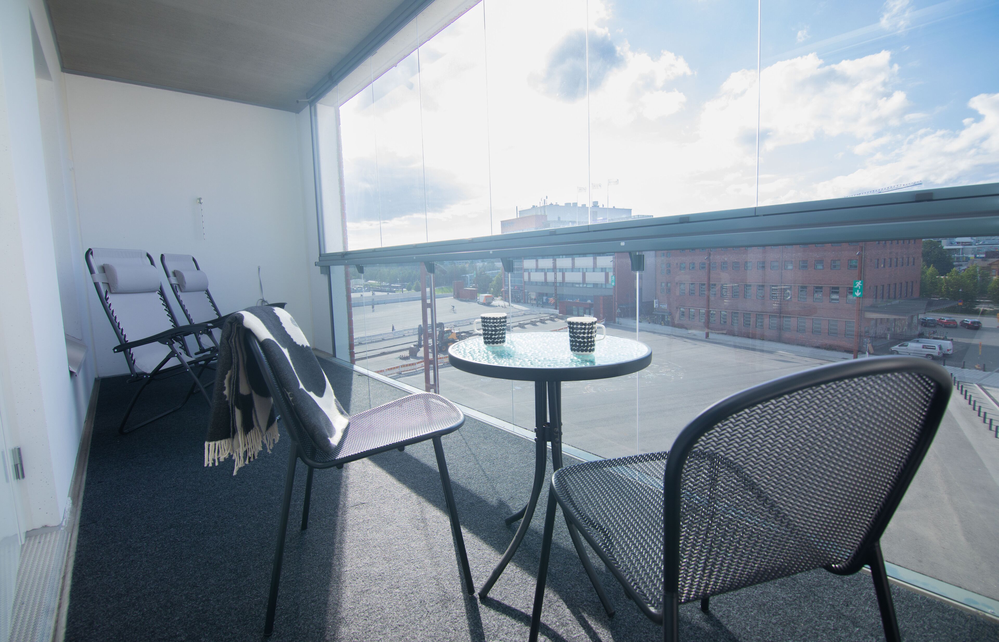 A balcony with a view of a cityscape, two lounge chairs, a small round table, and a chair.