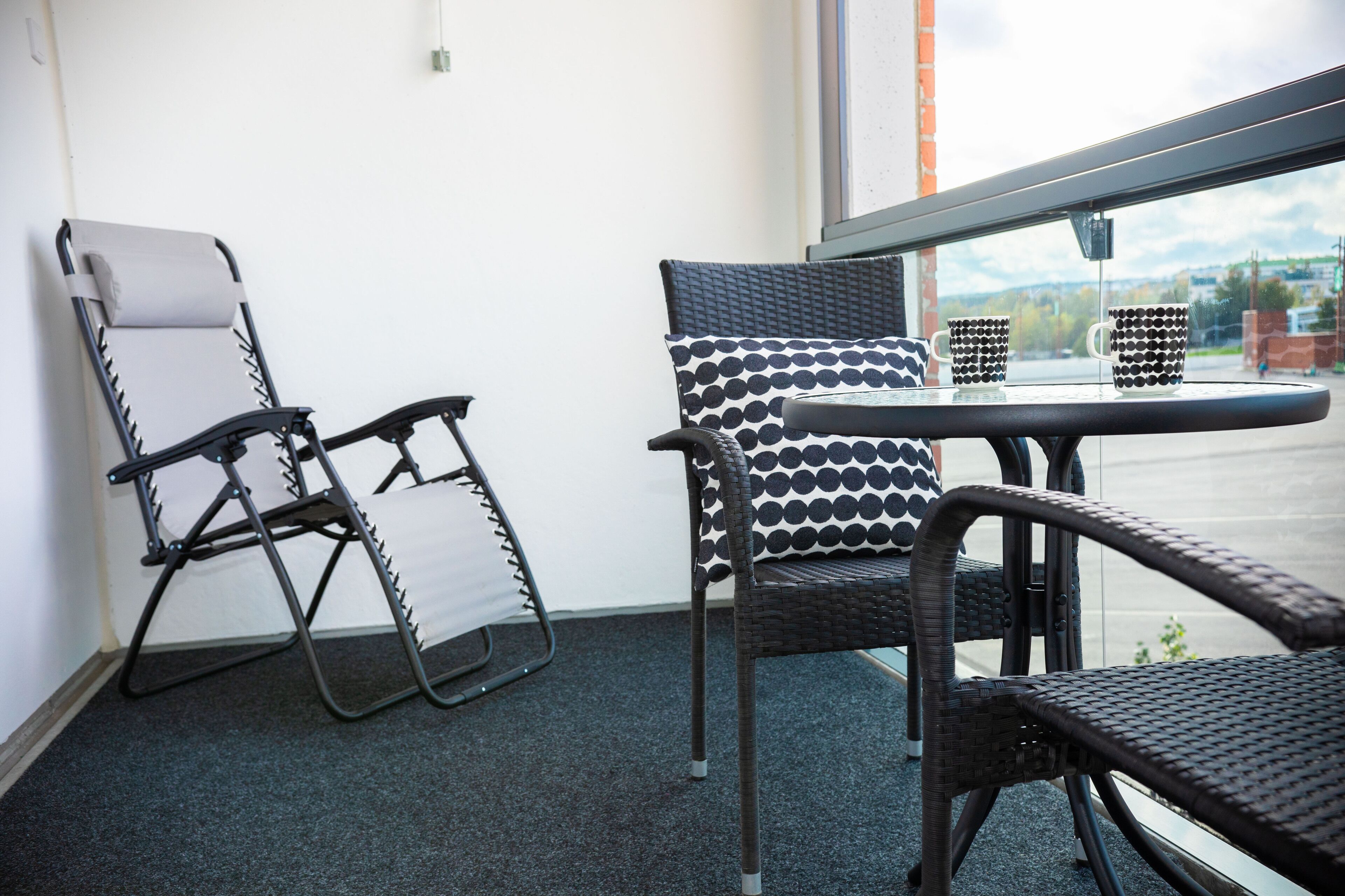 A corner of a room with a folding chair, a wicker chair, and a small table with two patterned mugs.