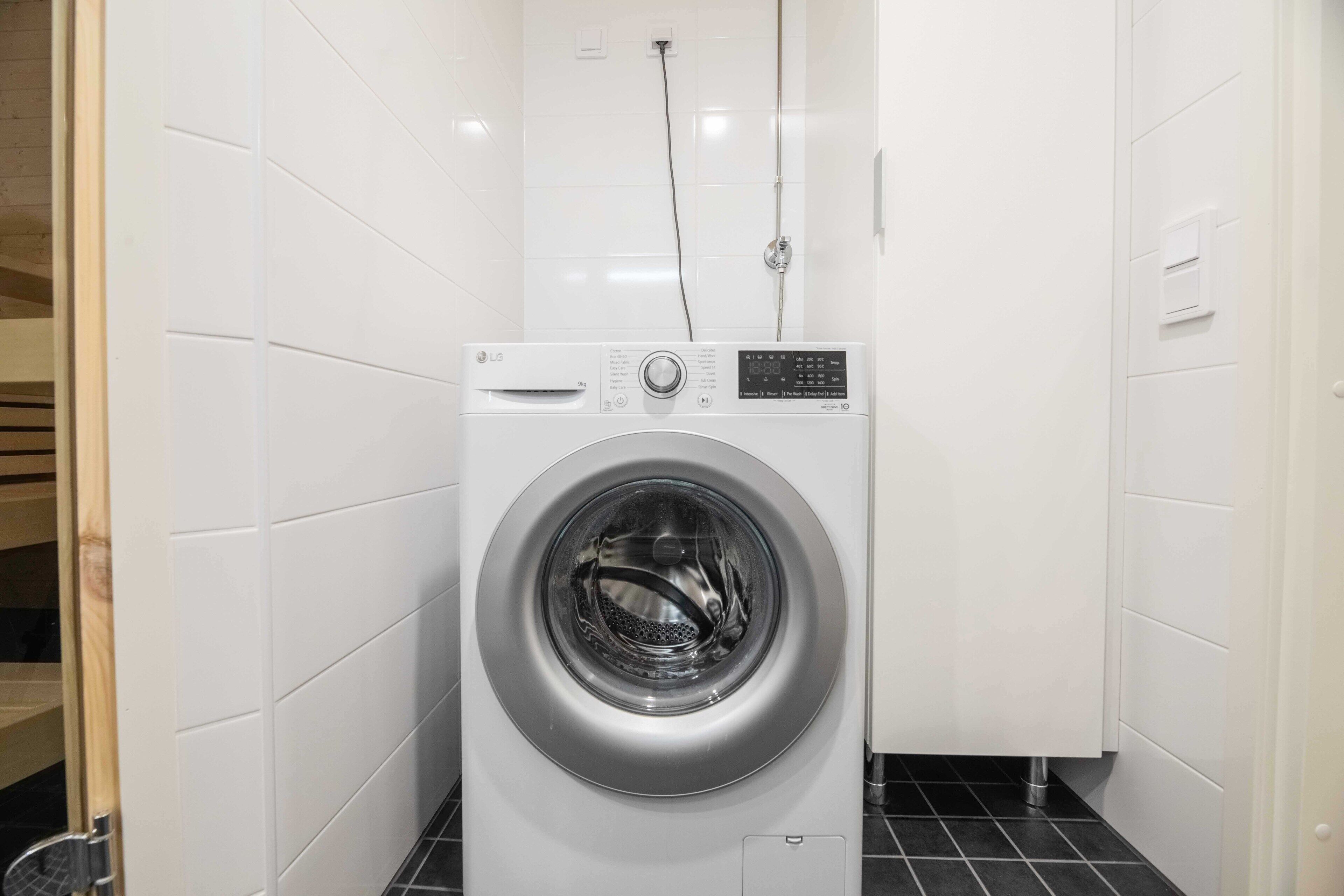Une petite buanderie moderne, dotée d’un lave-linge à chargement frontal, d’un petit meuble de rangement et d’un sol carrelé.