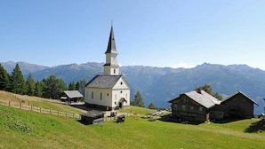 Une église dotée d’un clocher imposant, un petit bâtiment attenant et une structure ressemblant à une grange, le tout situé sur une colline verdoyante avec des montagnes en arrière-plan.
