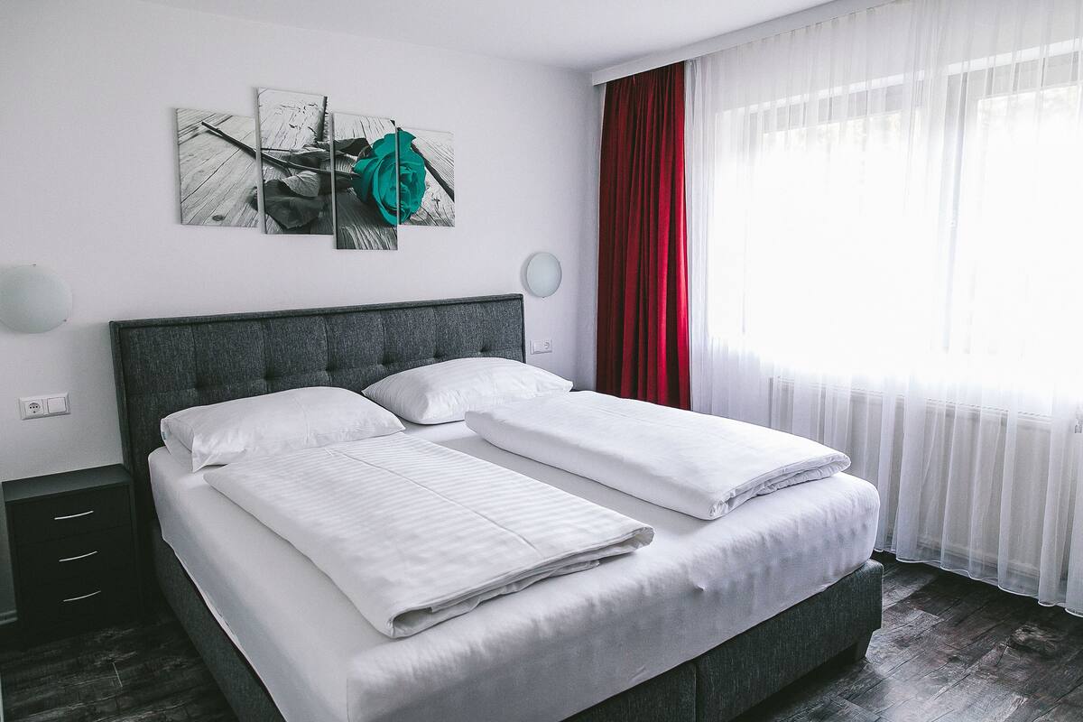 Una cama doble con ropa de cama blanca, un cabecero tapizado en gris y una mesita de noche con una lámpara.