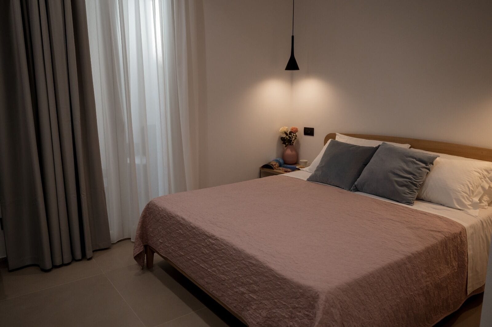 Una cama bien tendida con una colcha rosa, un cabecero de madera y una mesita de noche con un florero.