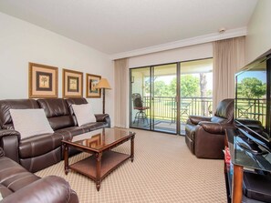 Ein Hotelzimmer mit braunem Ledersofa, einem Holztisch, einem Klavier und einem Balkon mit Blick auf Grünflächen.
