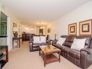 A living room with a brown leather sofa, a wooden coffee table, and a kitchen in the background. - Tranquil Saddlebrook 2bd/2ba Condo W/Views (Wesley Chapel)