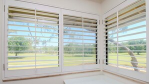 Una habitación con un escritorio en esquina y ventanas grandes con persianas horizontales, que ofrece vistas a un paisaje verde.