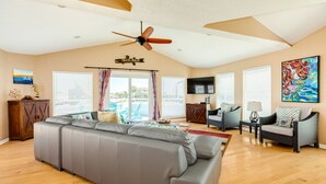 Ein geräumiges Wohnzimmer mit einem großen, grauen Ecksofa, einem Deckenventilator und Blick ins Freie durch große Fenster.