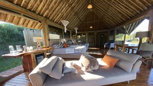 A cozy, well-lit room with a wooden ceiling and a large sofa with cushions. There is a wooden table with a coffee maker and cups, and a large window providing natural light. - A perfect mix of uncluttered safari simplicity, muted khaki tones. (Talek)
