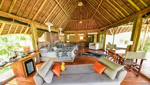 A spacious, well-lit room with a high wooden ceiling, a sofa, a dining table, and a television. - A perfect mix of uncluttered safari simplicity, muted khaki tones. (Talek)