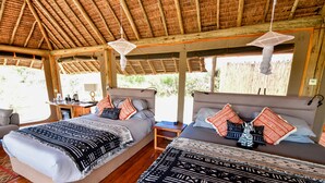 A room with two beds, each with patterned bedding and pillows, a wooden desk with a lamp, and a view of greenery outside. - A perfect mix of uncluttered safari simplicity, muted khaki tones. (Talek)