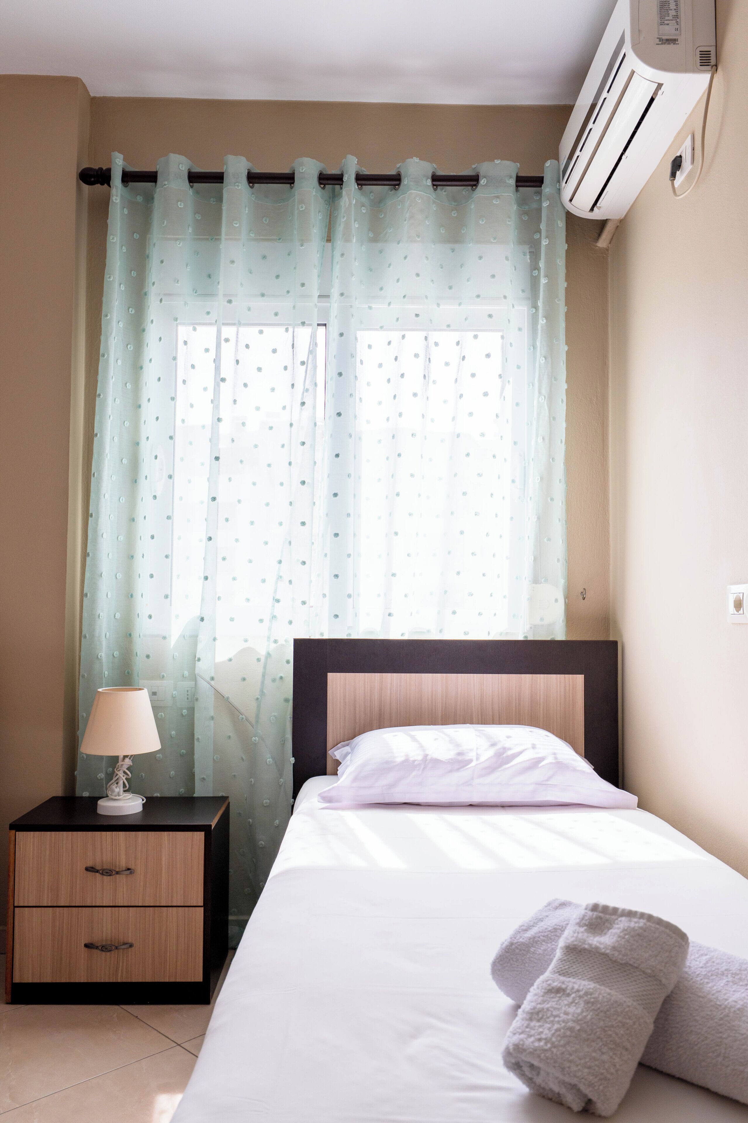 A neatly made bed with white linens and a rolled towel, a bedside lamp, a small wooden nightstand with drawers, and a window with sheer curtains.