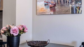 A dining table with a vase of flowers, a woven basket, and a painting of a harbor scene.