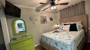 A bedroom with a bed, a ceiling fan, a TV, a green dresser, and a wall with decorative items.