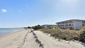 A beachfront hotel with a sandy walkway leading to the ocean, a multi-story building with balconies, and a clear sky.