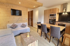 Una sala de estar moderna con un sofá, una mesa de centro y un comedor con mesa y sillas. Hay un televisor montado en la pared, un microondas y una estufa en la cocina.