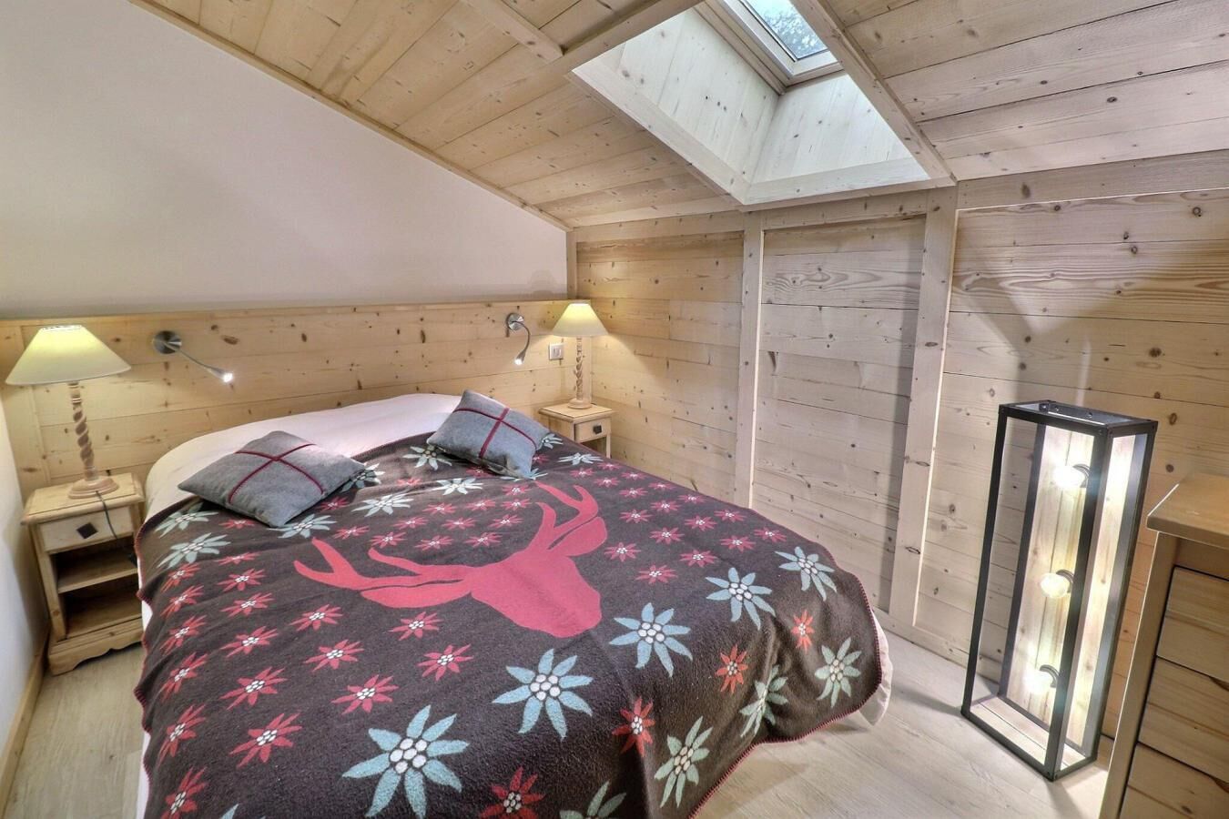 Une chambre confortable avec un plafond en bois, une lucarne, un lit avec une literie à motifs floraux, des lampes de chevet et une lampe sur pied.