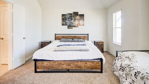 Un dormitorio con estructura de cama de madera, colcha blanca y edredón floral gris y blanco. Hay una obra de arte en la pared sobre la cama y una ventana con persianas a la derecha.