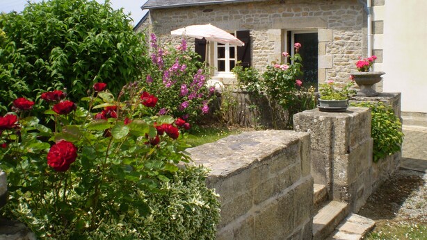 Une maison en pierre avec un toit en pente, un petit porche agrémenté d’un parasol blanc et un jardin où poussent des fleurs rouges et roses.