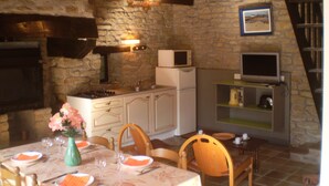 Une cuisine rustique aux murs de pierre, avec des poutres en bois, une table à manger dressée avec des serviettes orange et un vase vert rempli de fleurs.