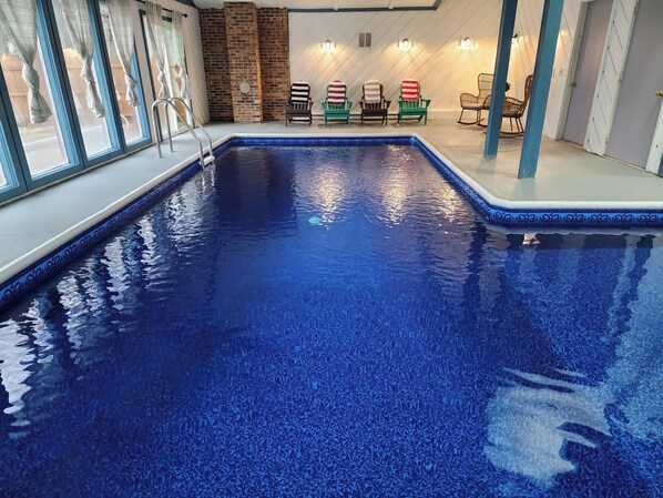 An indoor swimming pool with a blue tiled floor, a brick wall, and several chairs with striped and solid-colored covers.