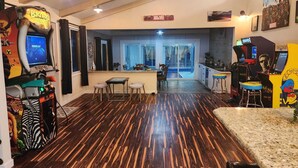 Una habitación con piso de madera, una zona de bar con taburetes, juegos de arcade y un comedor con sillas y una mesa.