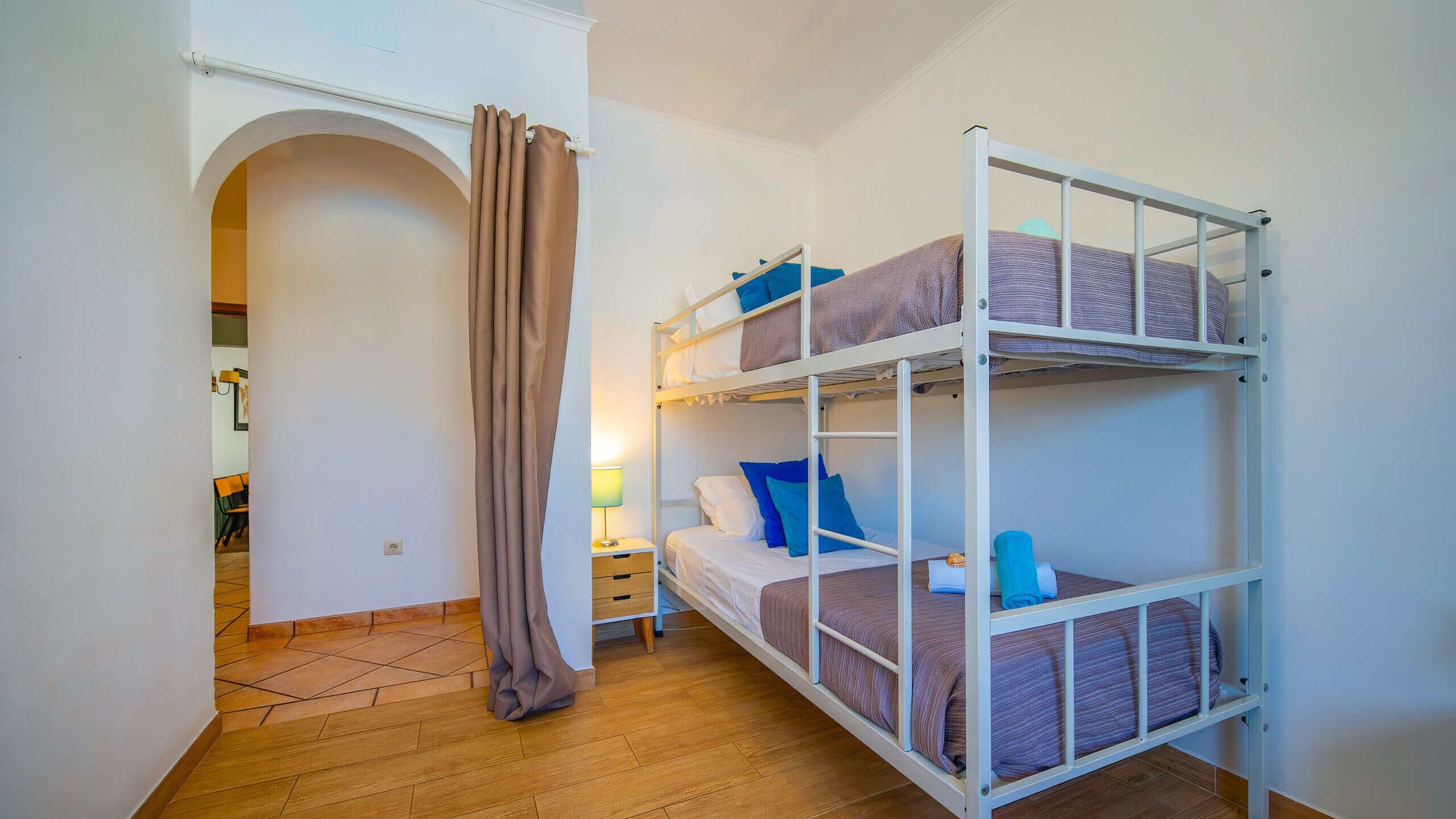 Ein Schlafraum mit Etagenbetten, Holzboden, Nachttisch und Lampe sowie einem Vorhang.