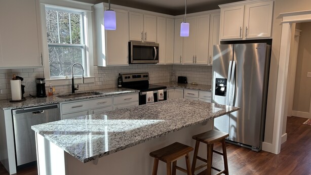 Una cocina moderna con armarios blancos, encimeras de granito, electrodomésticos de acero inoxidable e iluminación colgante.