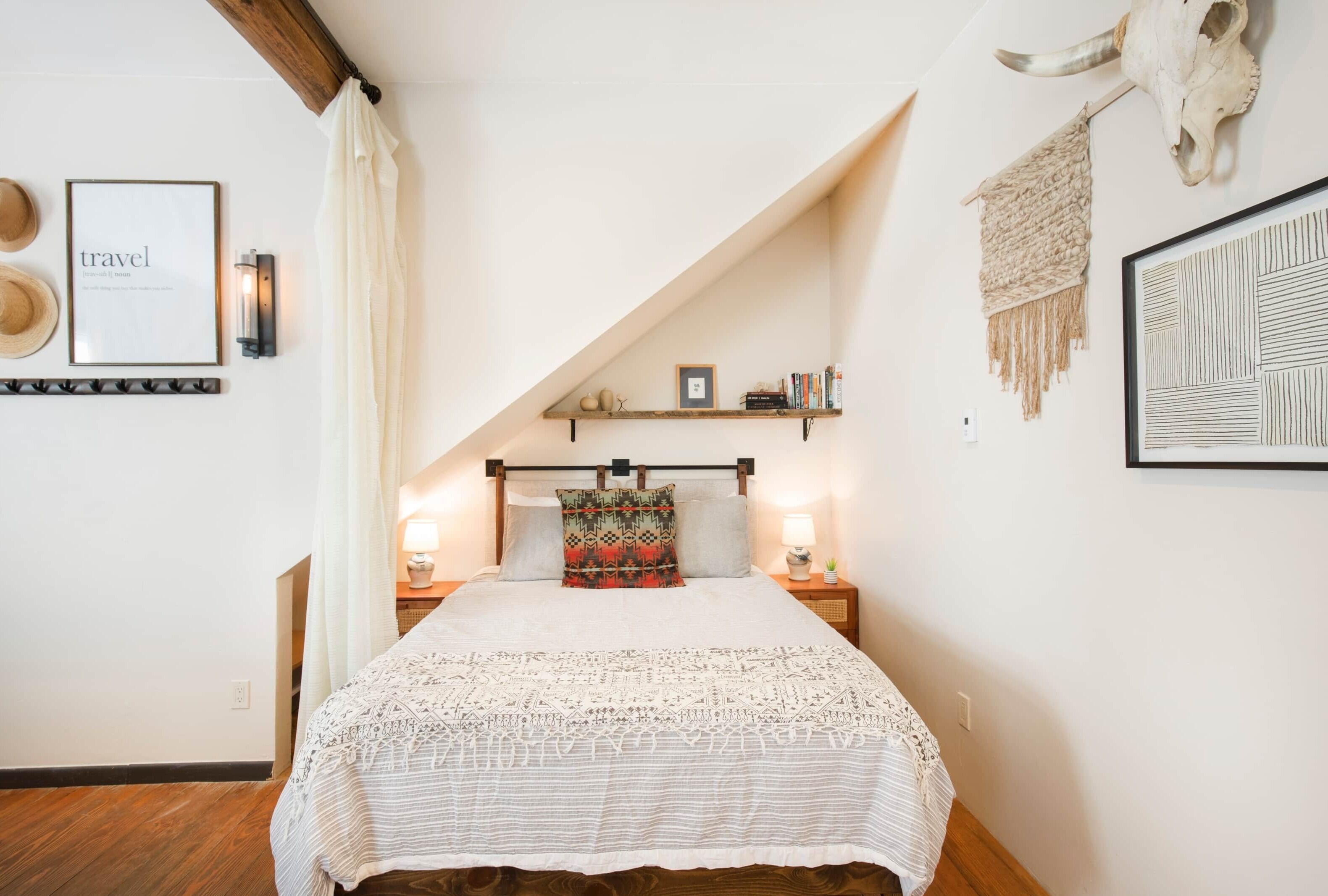 Une chambre avec un plafond en pente, un lit recouvert d’une couvre-lit à motifs, une étagère murale ornée de livres et d’objets décoratifs, et un tableau encadré accroché au mur.