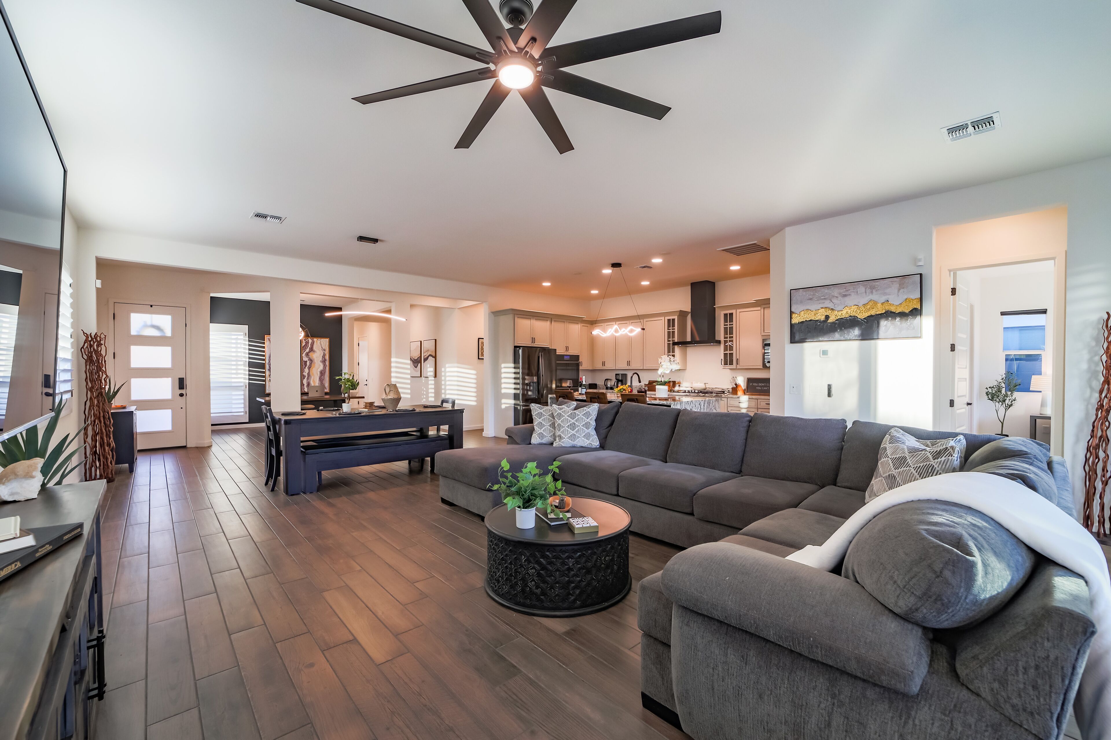 A spacious living room with a dark wood floor, a large gray sectional sofa, a coffee table with decorative items, and a ceiling fan.
