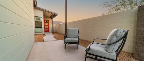 A patio area with two chairs and a concrete floor, adjacent to a building with a door and a window.